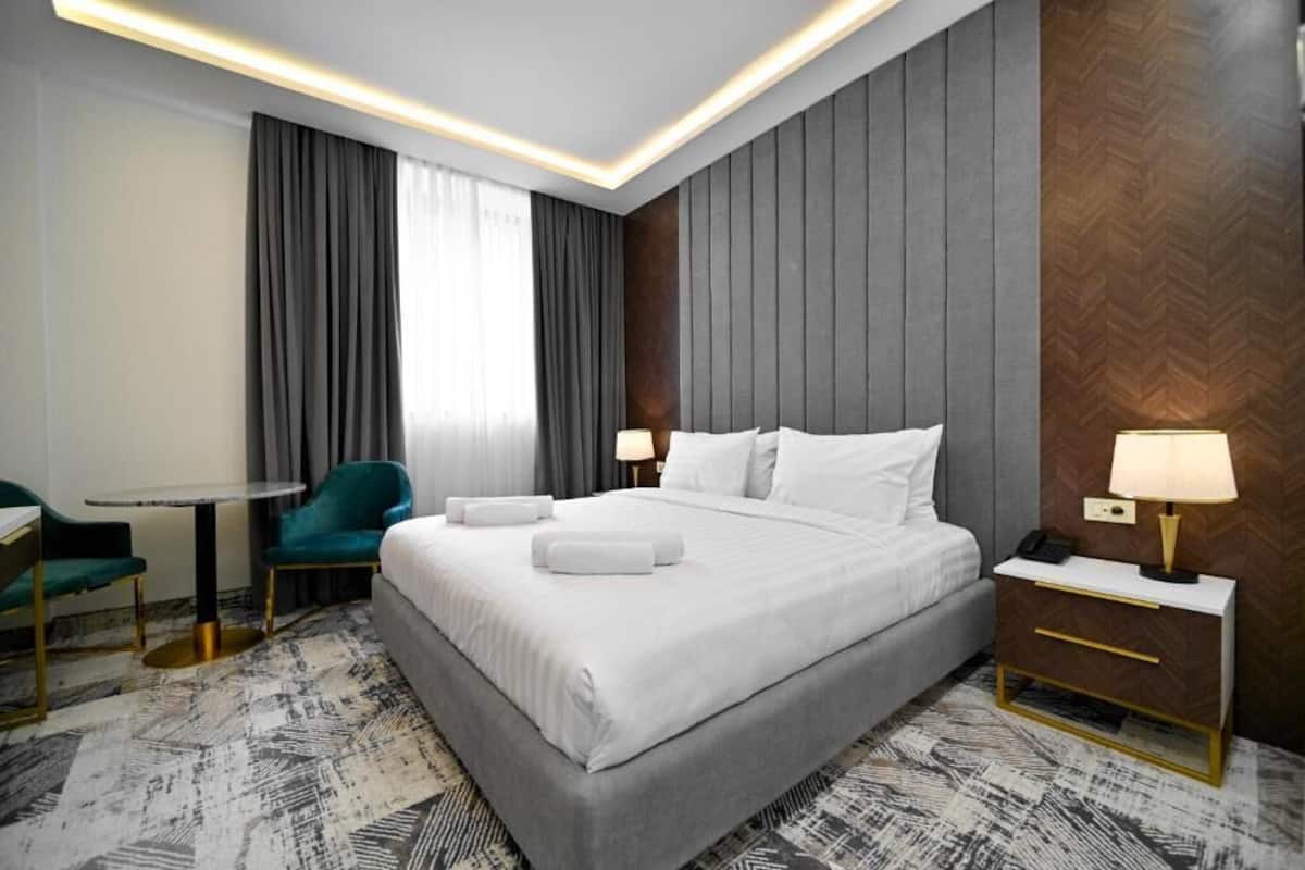 A modern hotel room with a large bed, a desk, a chair, and a nightstand with a lamp.