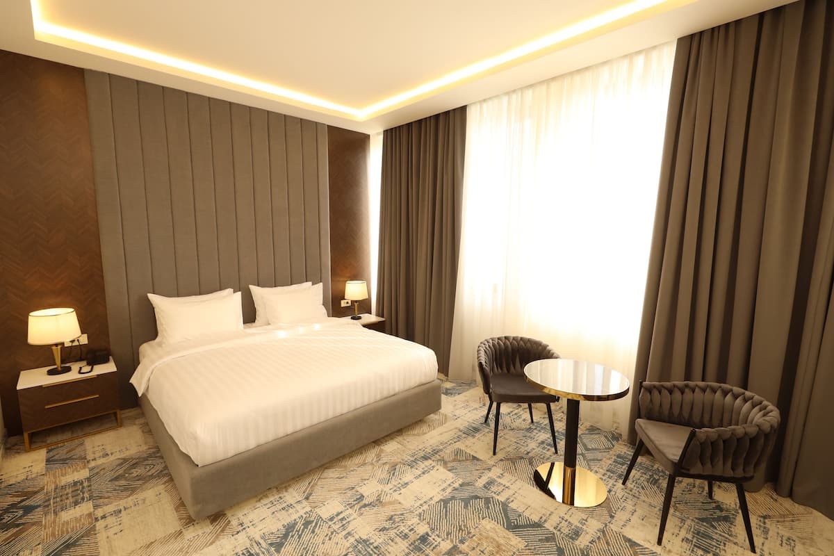 A hotel room with a large bed, two bedside tables with lamps, a small round table with a chair, and a view of the outdoors through curtains.