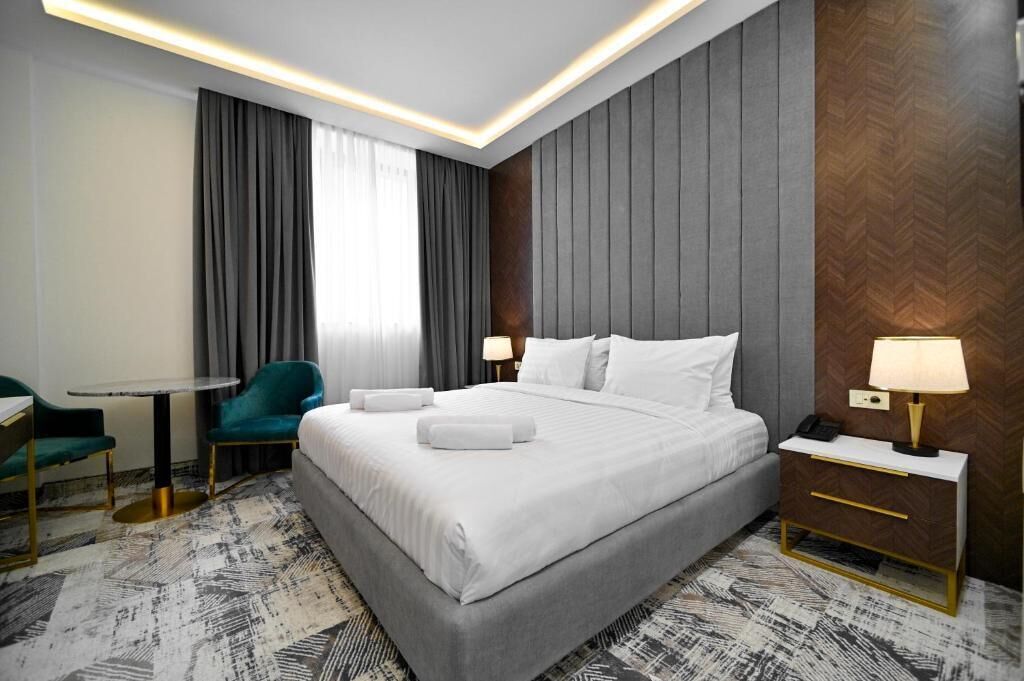 A modern hotel room with a large bed, a desk, a chair, and a nightstand with a lamp.
