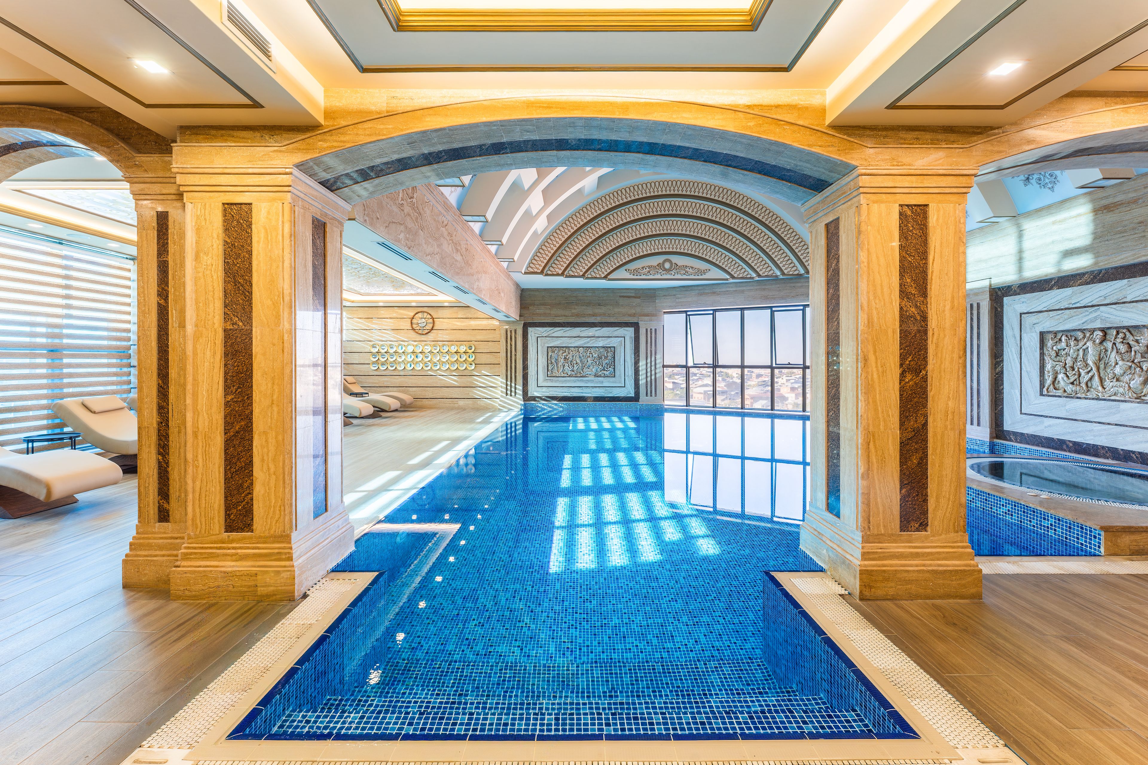 En luksuriøs indendørs swimmingpool med flisegulv, omgivet af marmorkolonner og et hvælvet loft med ovenlys.