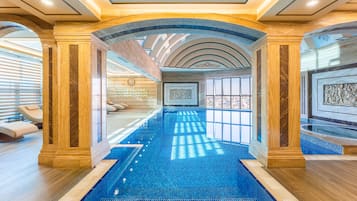 En luksuriøs indendørs swimmingpool med flisegulv, omgivet af marmorkolonner og et hvælvet loft med ovenlys.
