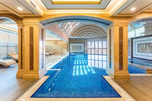 A luxurious indoor swimming pool with a tiled floor, surrounded by marble columns and a high ceiling with skylights.