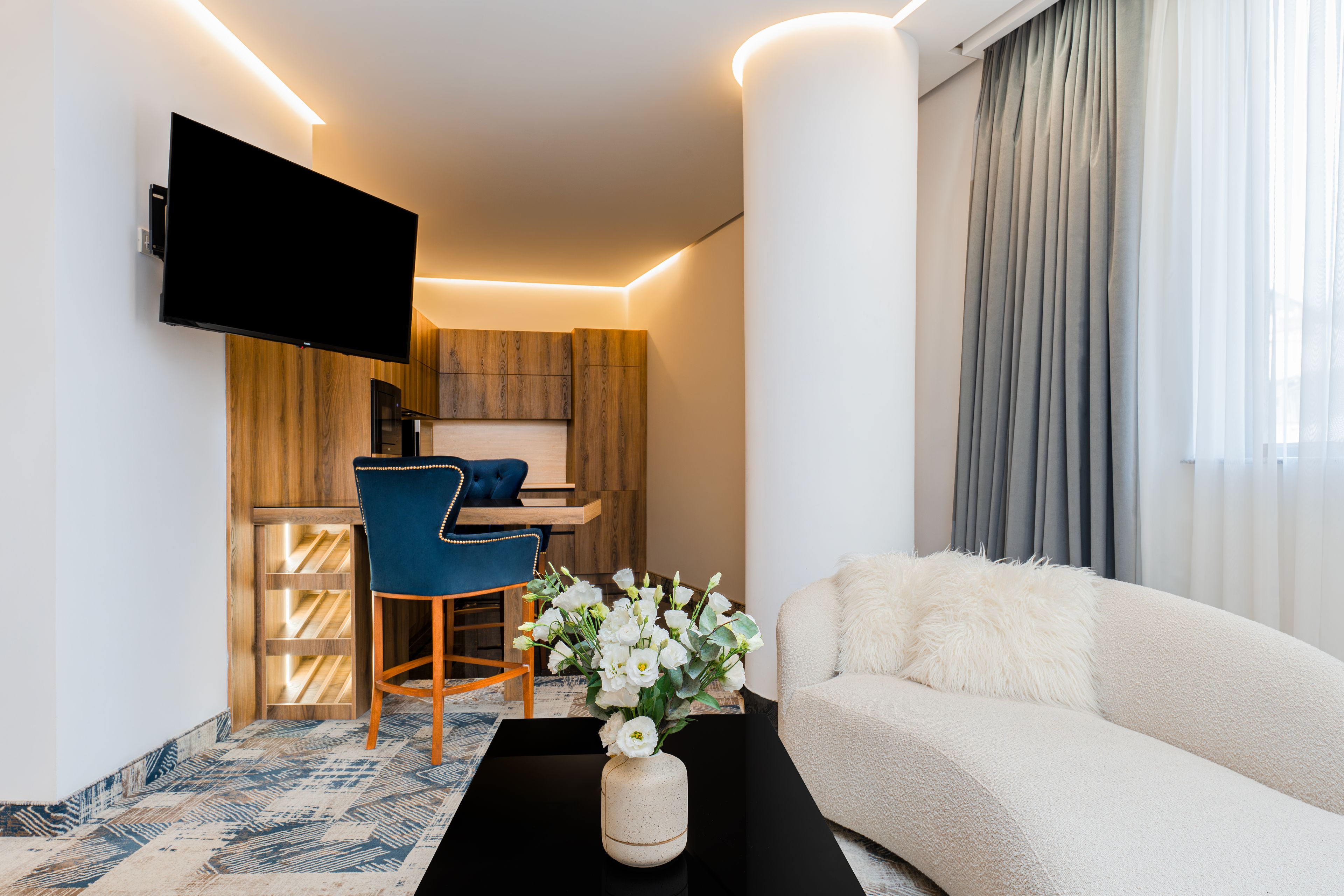 A modern living room with a flat-screen TV mounted on the wall, a sofa, a coffee table with a vase of flowers, and a dining area with a chair and wooden cabinets.