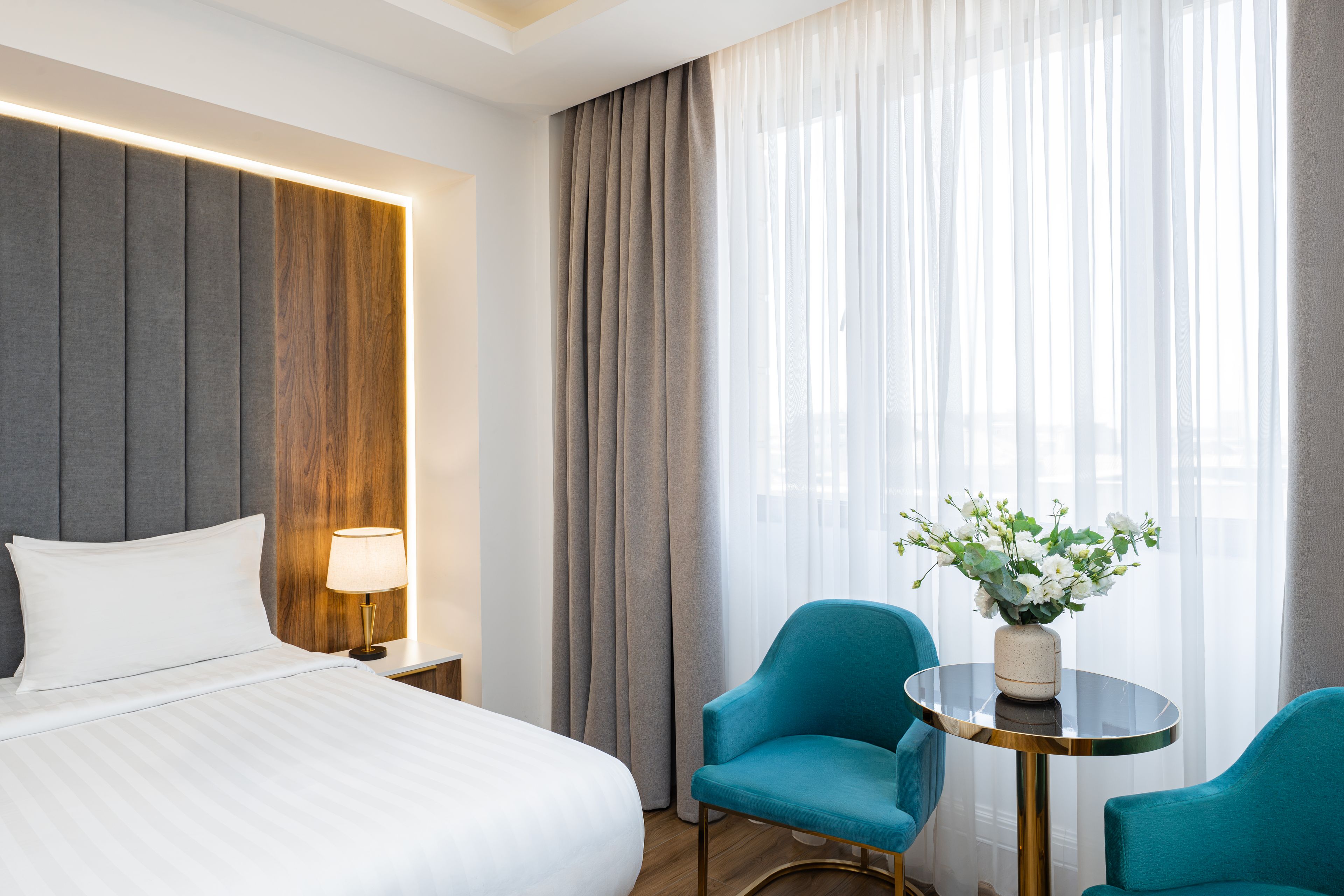 Habitación de hotel con cama, mesita de noche, lámpara, mesa con sillas y ventana con cortinas.