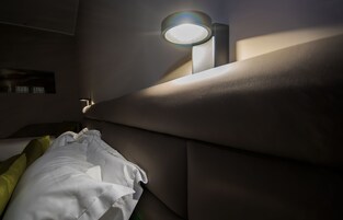 Hämärästi valaistu huone, jossa on sänky, tyyny ja seinään kiinnitetty valaisin.