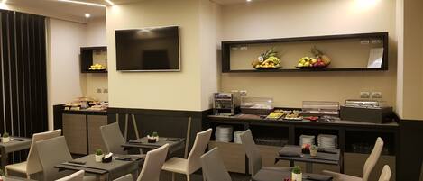 Un comedor moderno con mesas y sillas, un bufé con frutas y bebidas, y un televisor de pantalla plana en la pared.