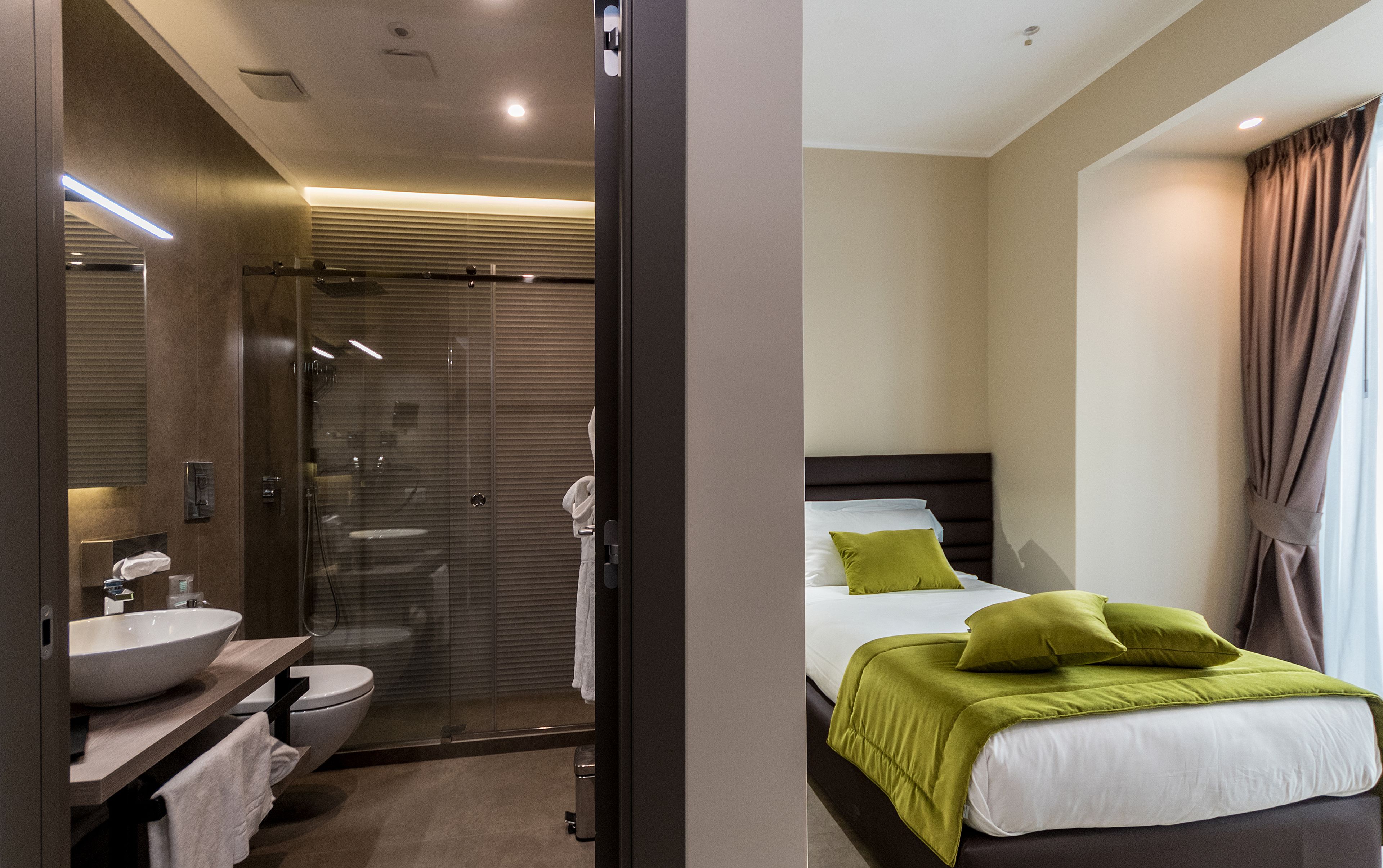 Una camera d'hotel moderna con bagno dotato di cabina doccia in vetro, lavandino e toilette. La zona notte presenta un letto con cuscini verdi e una grande finestra con tende.