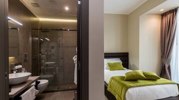 Una camera d'hotel moderna con bagno dotato di cabina doccia in vetro, lavandino e toilette. La zona notte presenta un letto con cuscini verdi e una grande finestra con tende.