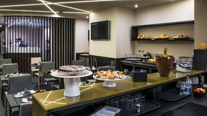 A modern hotel room with a dining area featuring a buffet with various pastries and a cake, a flat-screen TV mounted on the wall, and a fruit display on the back wall. - Hotel Matilde - Lifestyle Hotel (Naples)