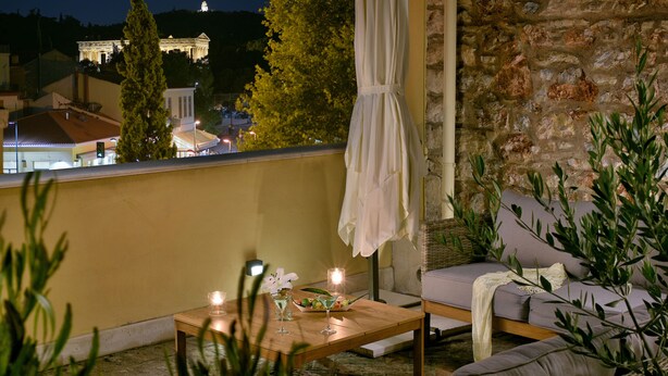 Un balcon avec un canapé en osier, une table en bois décorée de bougies et une vue sur un bâtiment de style classique illuminé la nuit.