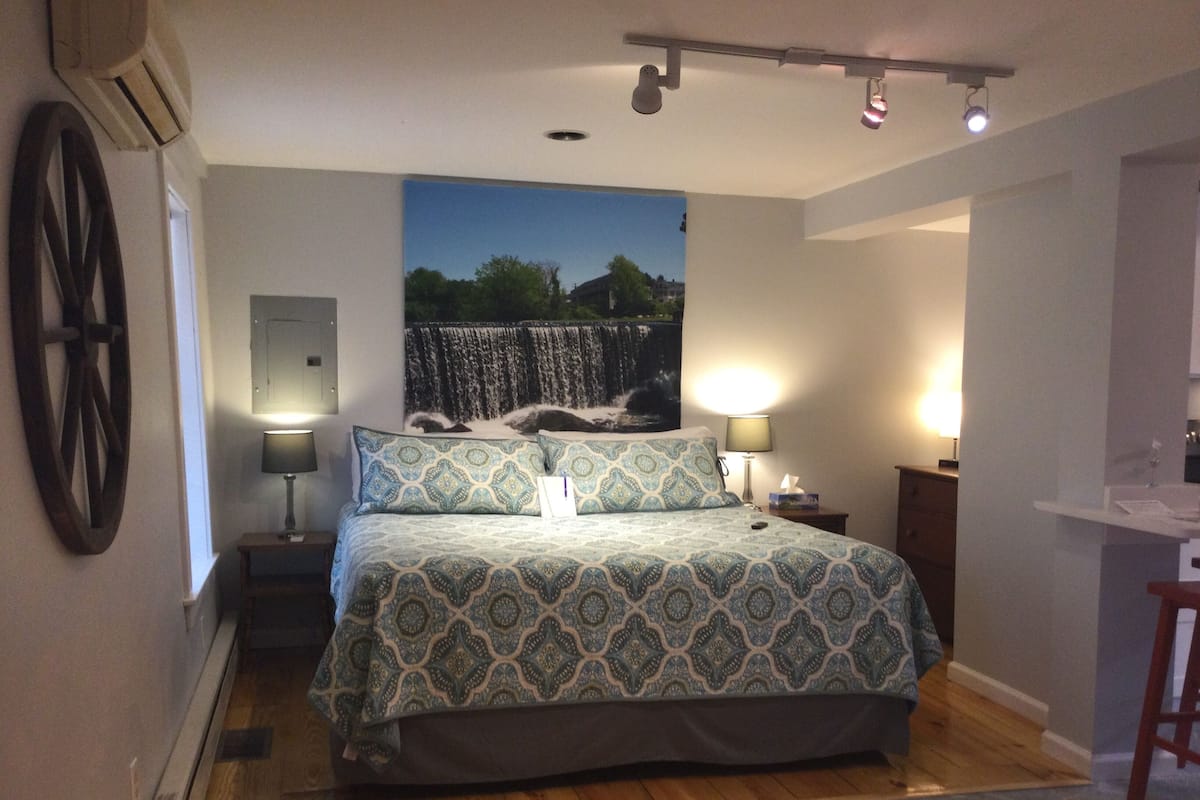 A bedroom with a bed, two bedside lamps, a wall-mounted air conditioner, a wooden floor, and a large wall tapestry.
