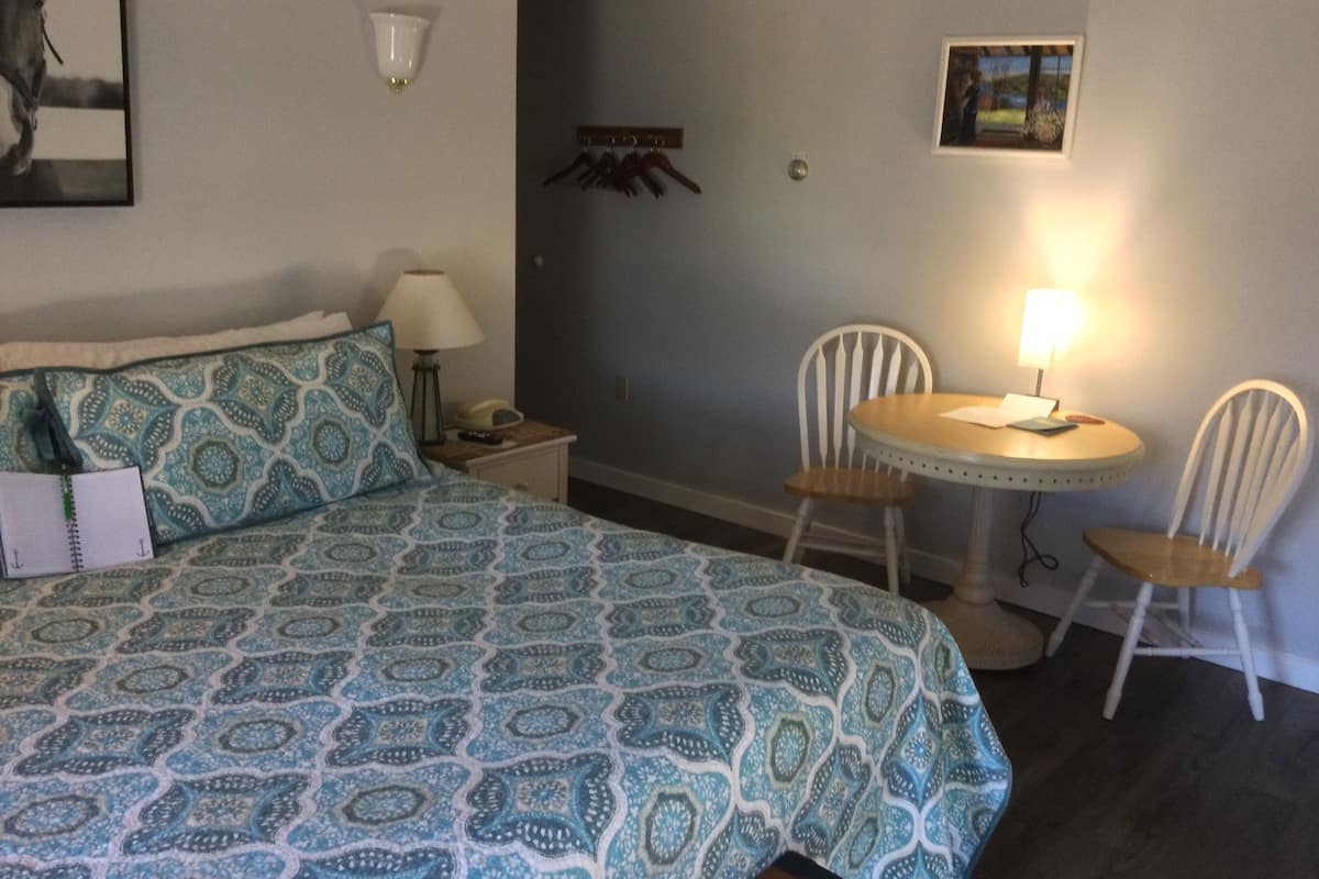 A hotel room with a bed, two chairs, a small table, a lamp, a nightstand, a wall-mounted lamp, a coat rack, and a framed picture.