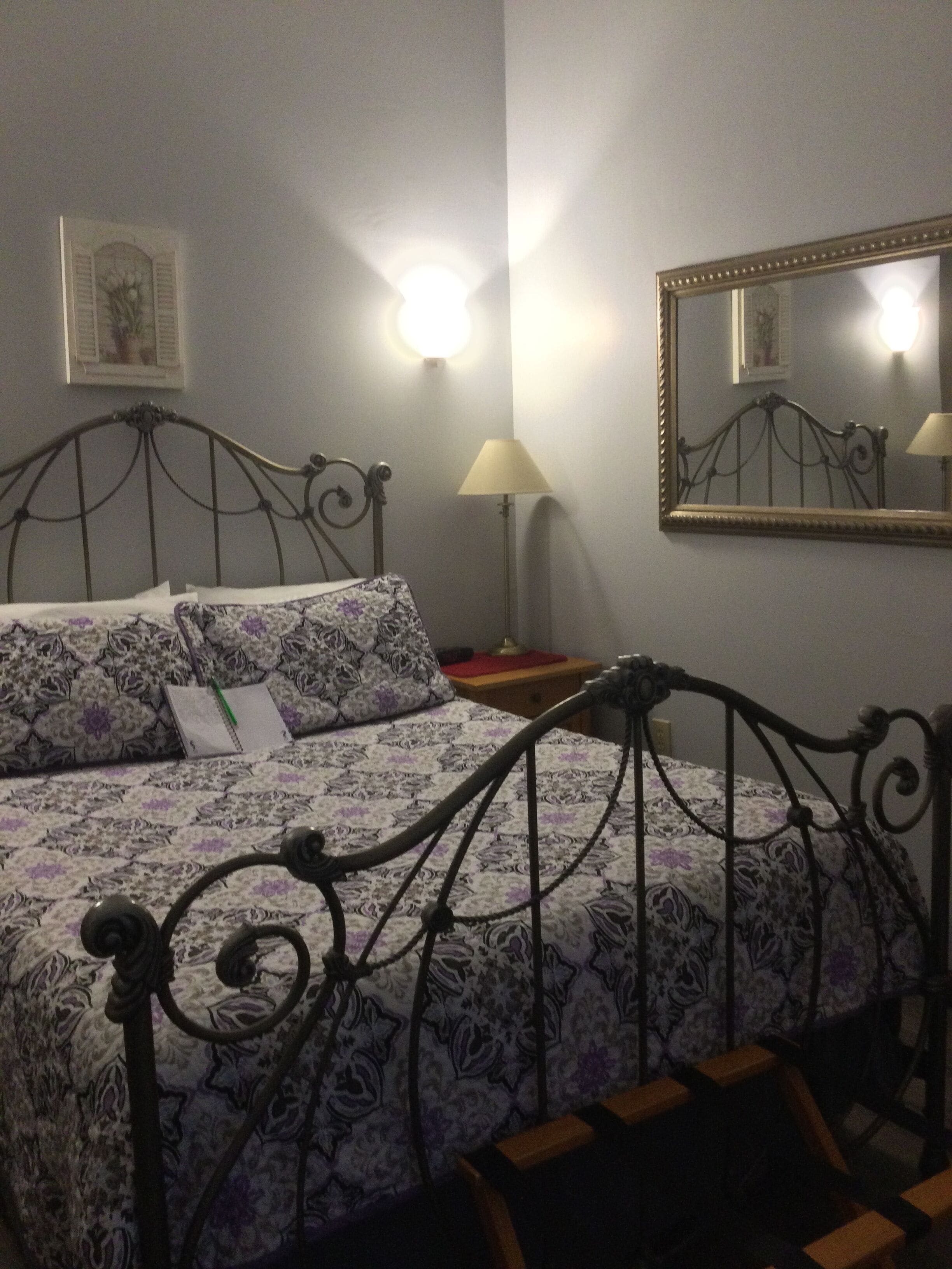 Ein ordentlich bezogenes Bett mit einem gemusterten Überwurf, ein Nachttisch mit einer Lampe und ein wandmontierter Spiegel.
