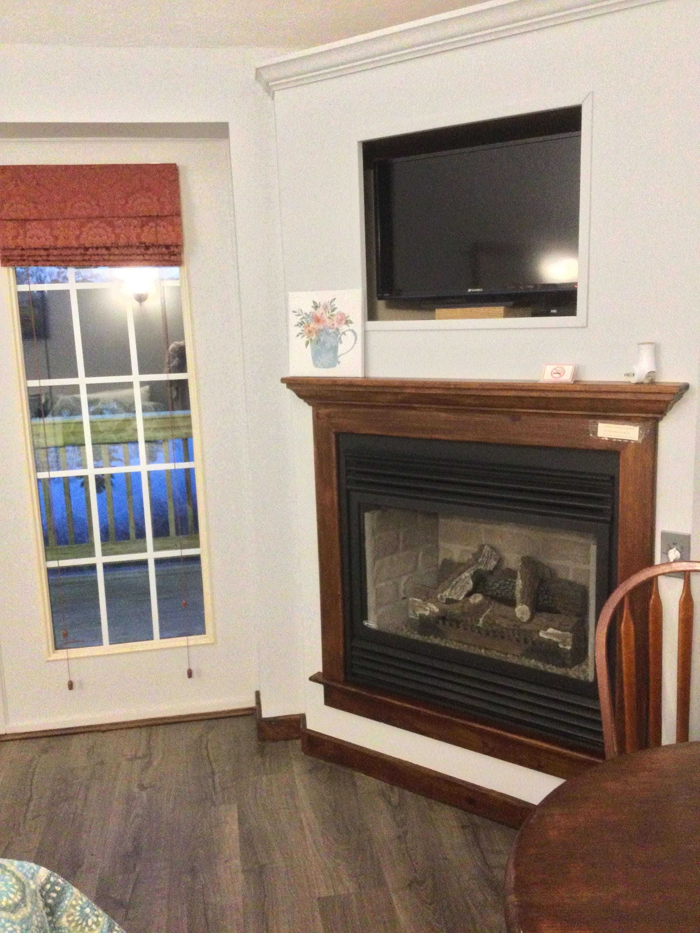 A living room with a fireplace, a television mounted above it, and a window with blinds.