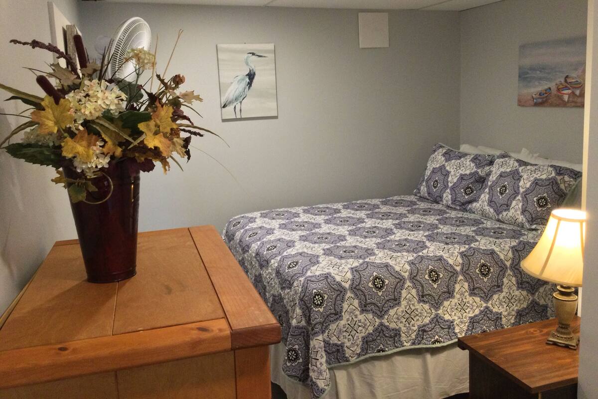 A bedroom with a bed, a wooden nightstand, a table with a vase of flowers, a wall fan, and a painting.