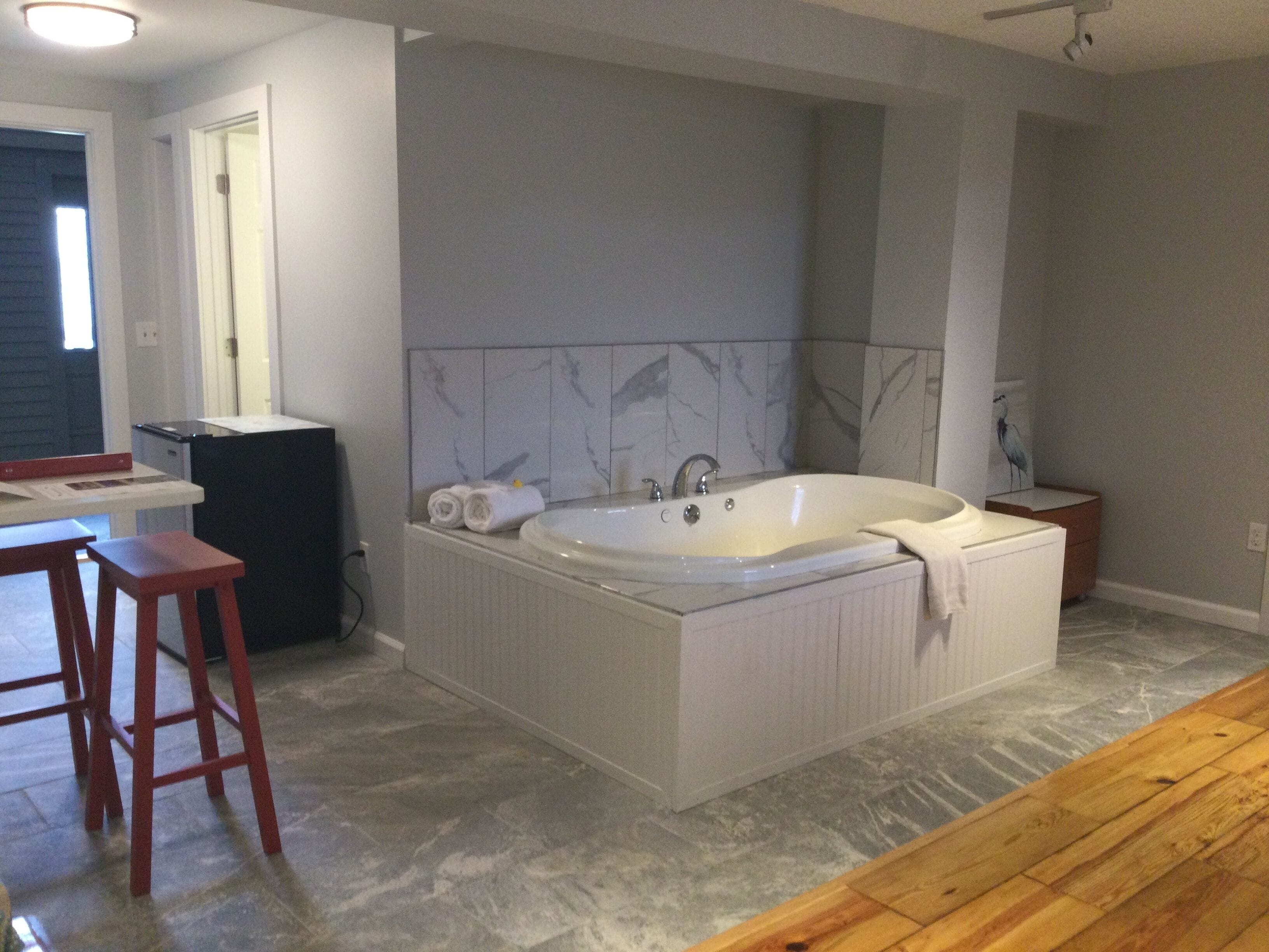 A bathroom with a large bathtub, a wooden stool, a small table, and a towel on the floor.