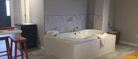 A bathroom with a large bathtub, a wooden stool, a small table, and a towel on the floor.