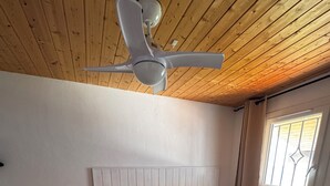 Un dormitorio con techo de madera, una cama con colcha estampada, dos almohadas azules, una mesita de noche con lámpara y una ventana con barrotes.