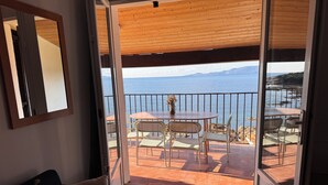 Habitación con puerta de vidrio corrediza que da a un balcón exterior con mesa y sillas, con vista a un cuerpo de agua y montañas.