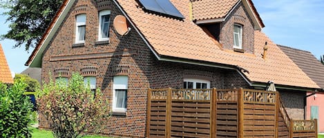 Uma casa de dois andares com fachada de tijolo, telhado inclinado e painéis solares, e cerca de madeira.