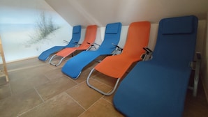 Une rangée de chaises longues avec des structures métalliques et des coussins bleus et orange, disposées dans une véranda dotée d’un sol carrelé et offrant une vue sur la plage.