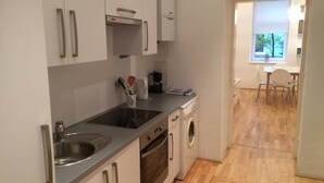 A compact kitchen with a sink, stove, and dishwasher, leading to a dining area with a table and chairs. - Modern, quiet apartment in an old building (Wien)