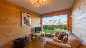 A cozy room with wooden walls, a large window offering a mountain view, a sofa, a television on a wooden table, and a bean bag. - Chalet Asphodel (Haute-Nendaz)