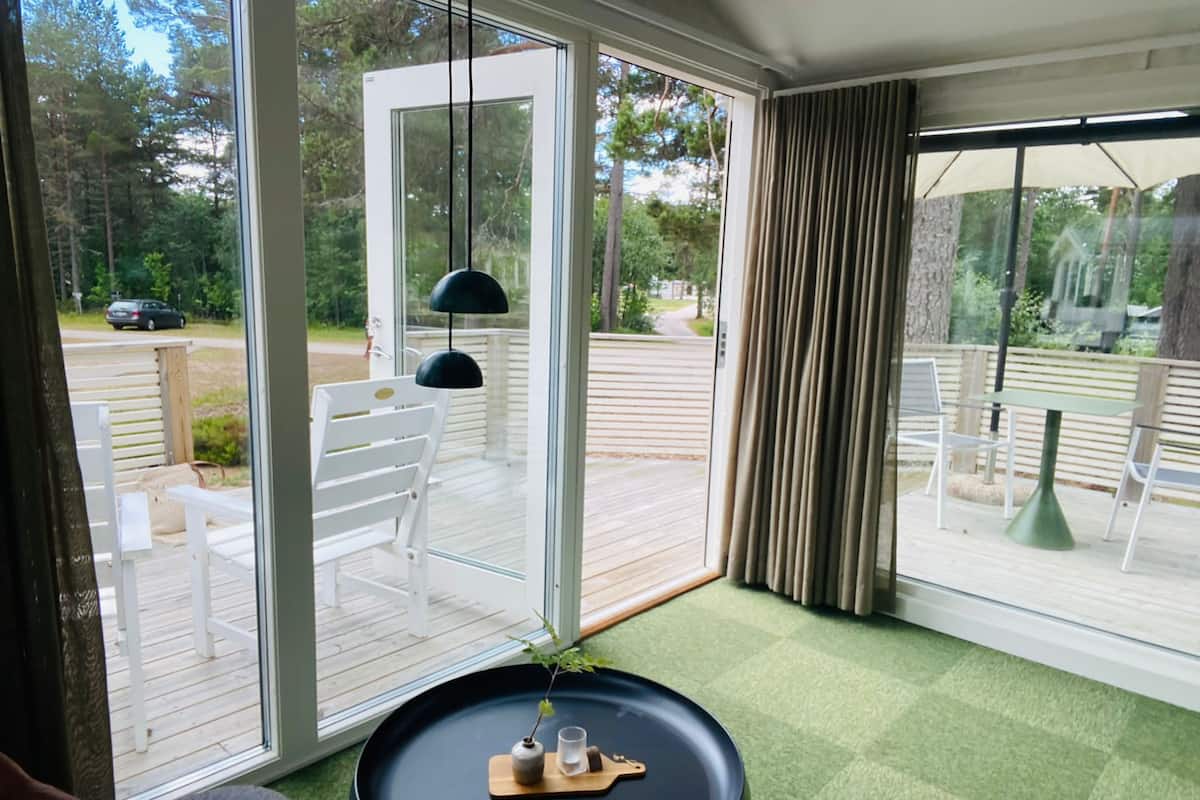 Ein Zimmer mit einer großen Schiebeglasdoor, die auf eine Terrasse im Freien führt. Dort steht ein runder Tisch mit einer Pflanze und einem Tablett, darüber schwebt eine schwarze Pendelleuchte.