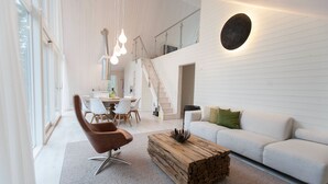 Uma sala de estar moderna com um sofá branco, uma poltrona marrom e uma mesa de centro de madeira. Há uma escada que leva a um andar superior, e uma área de jantar com mesa e cadeiras.