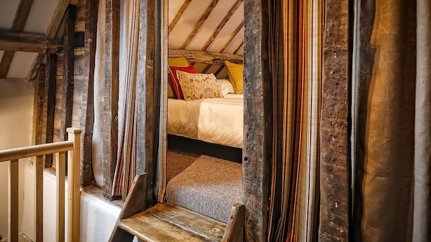 Eine rustikale Holztreppe führt zu einem Schlafzimmer auf dem Dachboden mit schrägem Dach und einem Bett mit bunten Kissen.