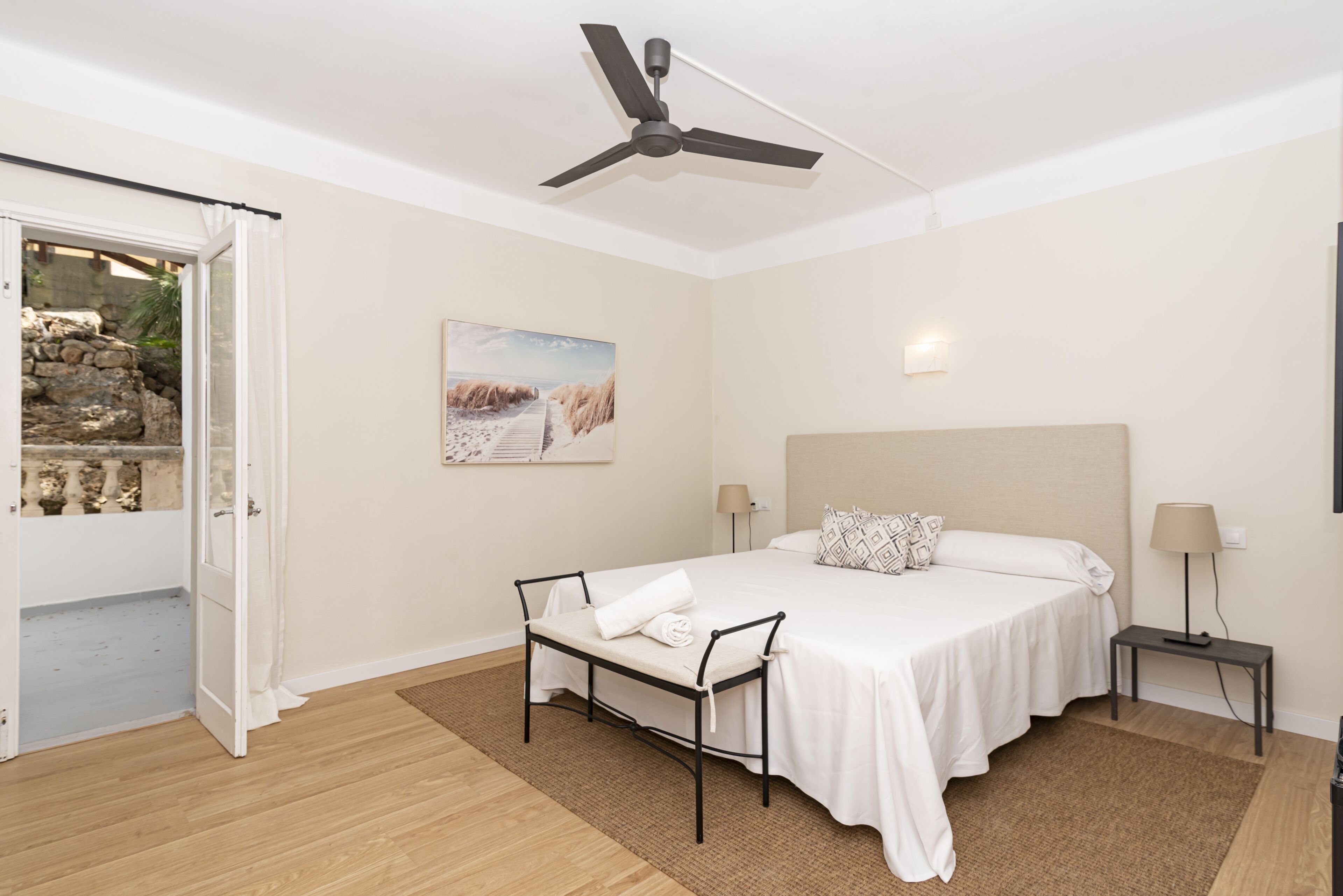 Un dormitorio con cama, ventilador de techo, mesita de noche con lámpara, ventana con cortinas y un cuadro en la pared.