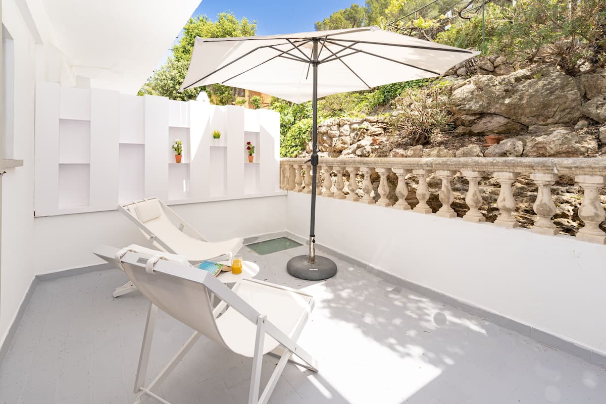 A white outdoor seating area with a table and chairs, a large umbrella, and a view of rocky terrain and greenery.