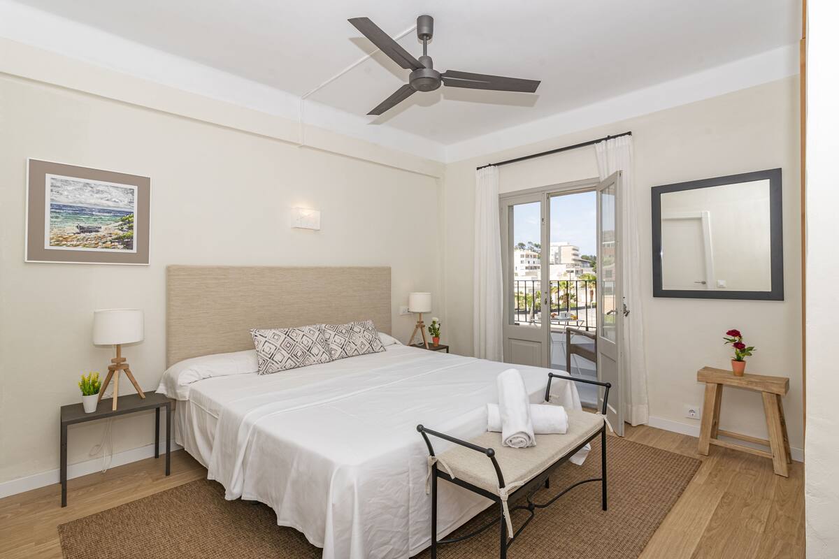 A hotel room with a bed, a ceiling fan, a window with curtains, a bedside table with a lamp, and a small bench.
