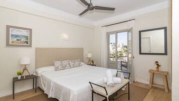 A hotel room with a bed, a ceiling fan, a window with curtains, a bedside table with a lamp, and a small bench.