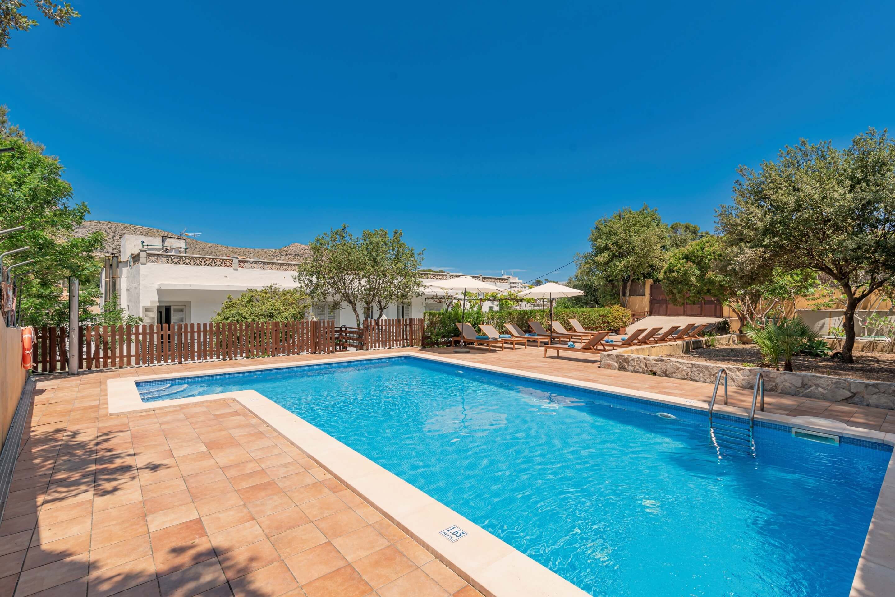 A rectangular swimming pool with a tiled deck, surrounded by lounge chairs and umbrellas, with a wooden fence and trees in the background.
