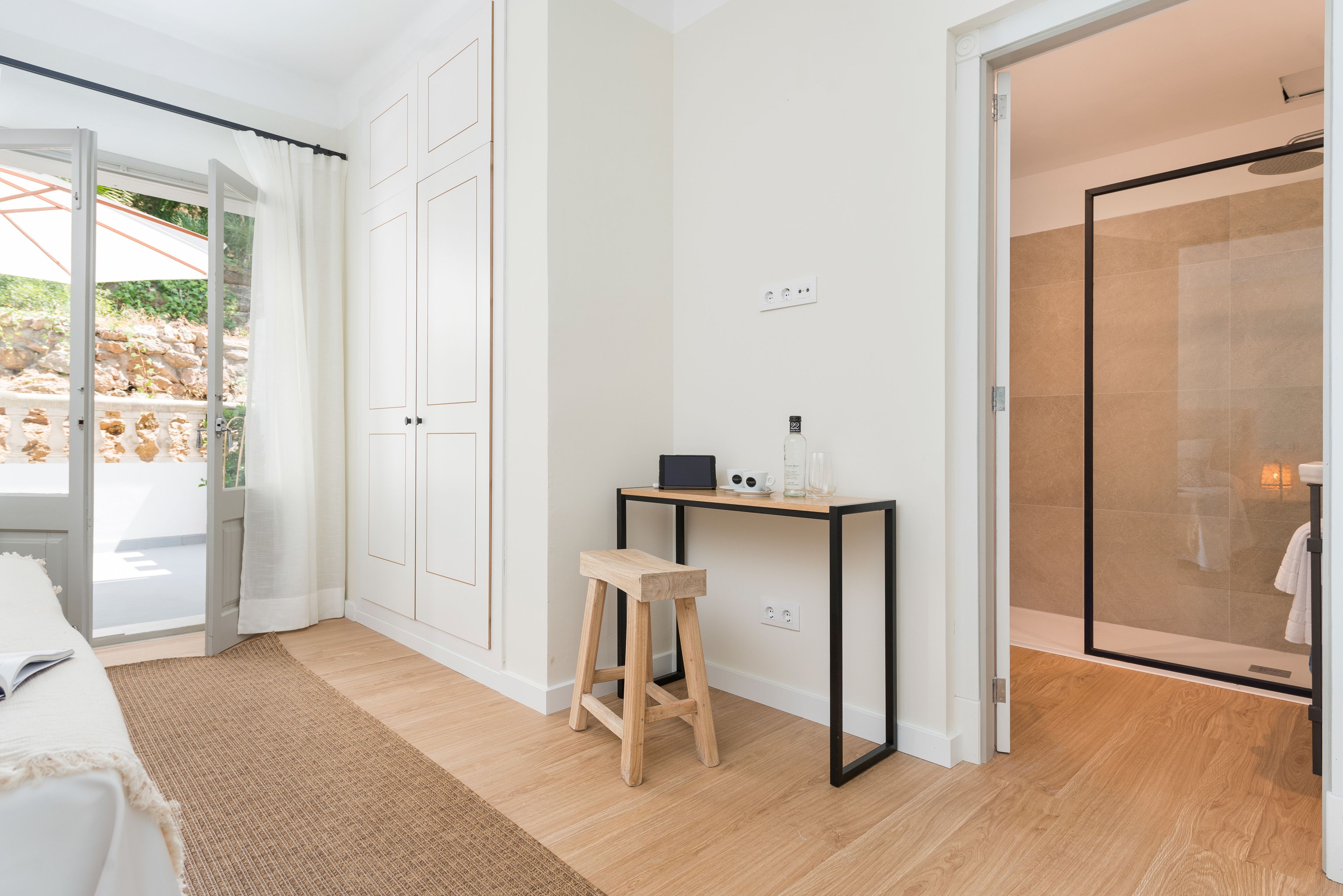 A modern room with a bed, a small wooden stool, a desk with a computer, and a glass shower.