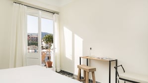 Une chambre d’hôtel avec un lit, un petit tabouret en bois, une table avec une chaise, et une vue sur les bâtiments et les montagnes par la fenêtre.