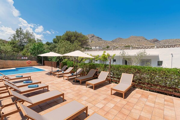 A poolside area with sun loungers and umbrellas, a swimming pool, and a view of a mountainous landscape.