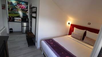 A bedroom with a large bed, a nightstand, and a wall-mounted light. There is a kitchenette with a microwave and a small fridge. A painting of a lotus flower is on the wall.