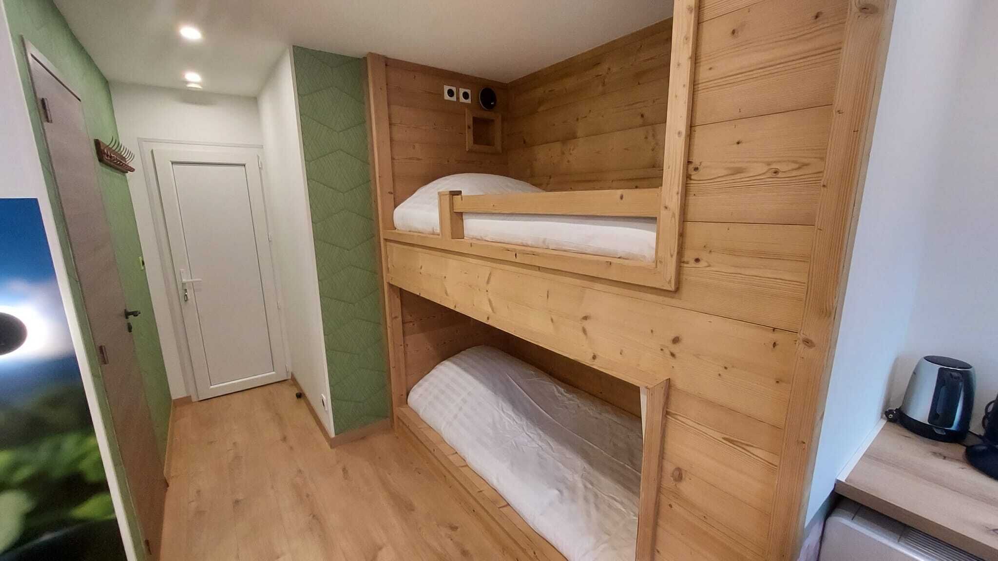 Une petite pièce avec un lit superposé, une petite fenêtre et une porte donnant sur une autre pièce.