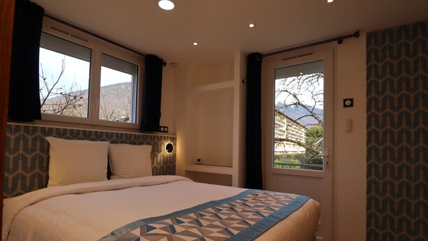 A bedroom with a large bed, a window with curtains, a door with a view, and patterned wallpaper. - Le Lotus Blanc (Albertville)