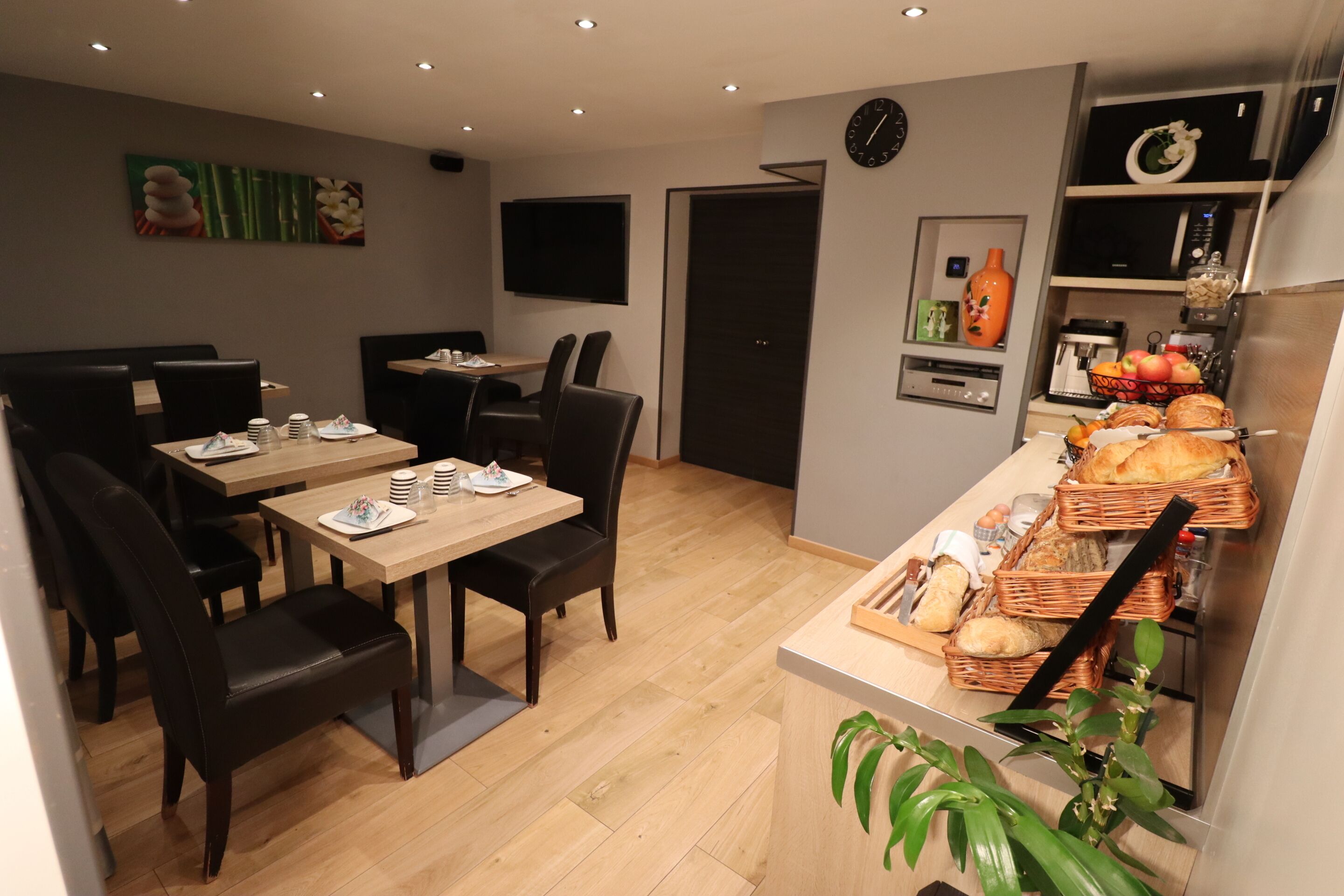 A modern dining area with wooden tables and chairs, a kitchen counter with bread and pastries, and a wall-mounted clock.
