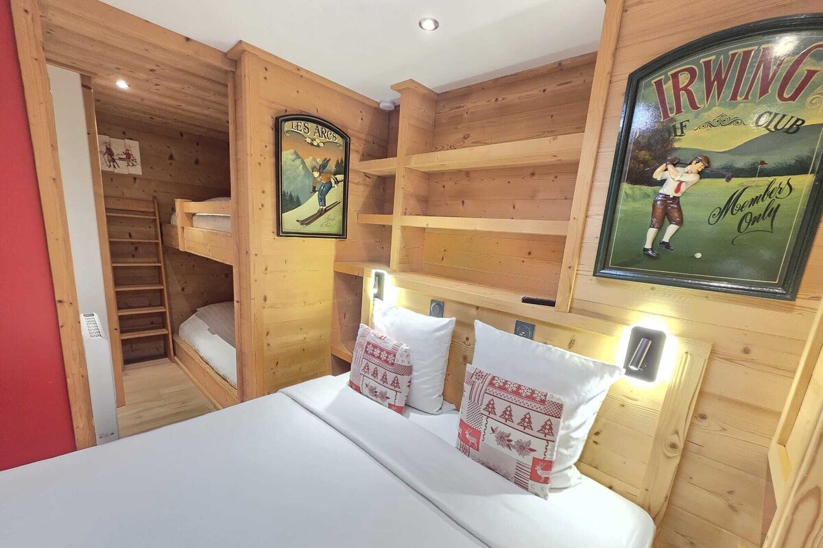 A wooden room with bunk beds, a bed with pillows, a framed poster, and a shelf.