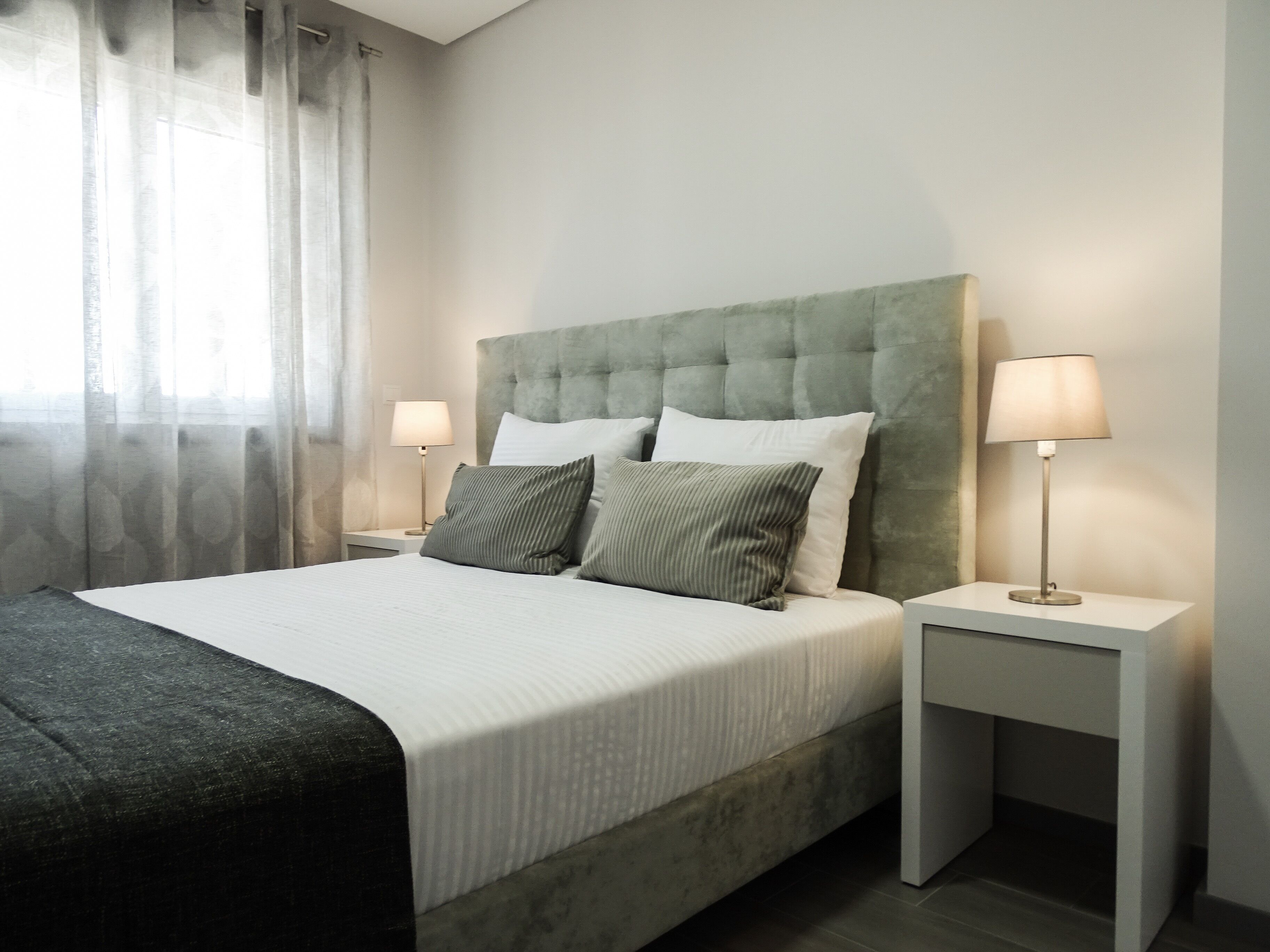 A modern bedroom with a tufted headboard, a large bed with white linens, two bedside tables with lamps, and sheer curtains.
