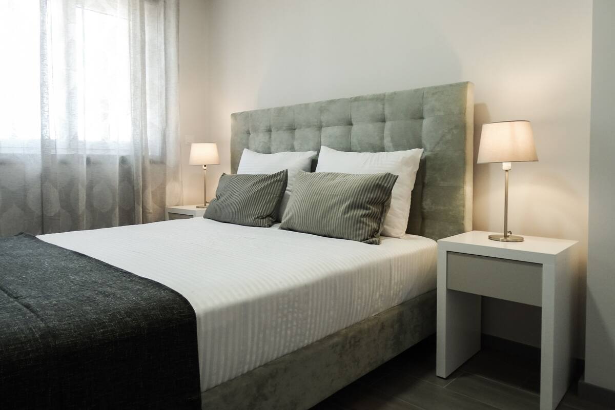 A neatly made bed with a grey headboard, two bedside tables with lamps, and a window with sheer curtains.