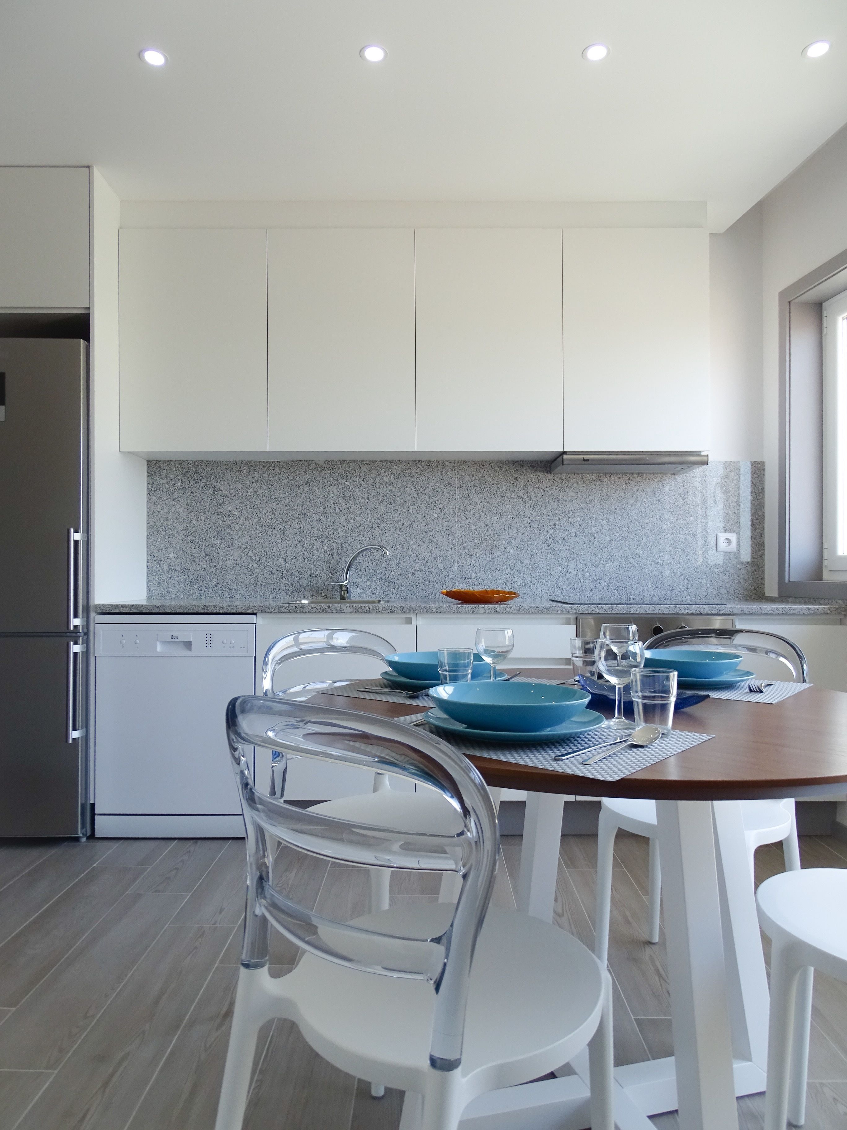 Une cuisine moderne avec des meubles blancs, un dosseret en granit et un coin repas comprenant une table en bois et des chaises blanches.
