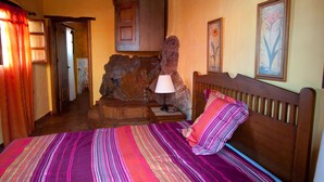 Ein Schlafzimmer mit Bett, Lampe, Holz-Kopfstück, Steinwand und einer Tür zu einem anderen Raum.