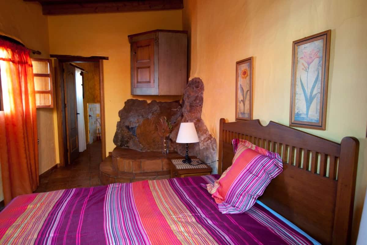 Une chambre à coucher comprenant un lit, une lampe, une tête de lit en bois, un mur en pierre et une porte donnant sur une autre pièce.