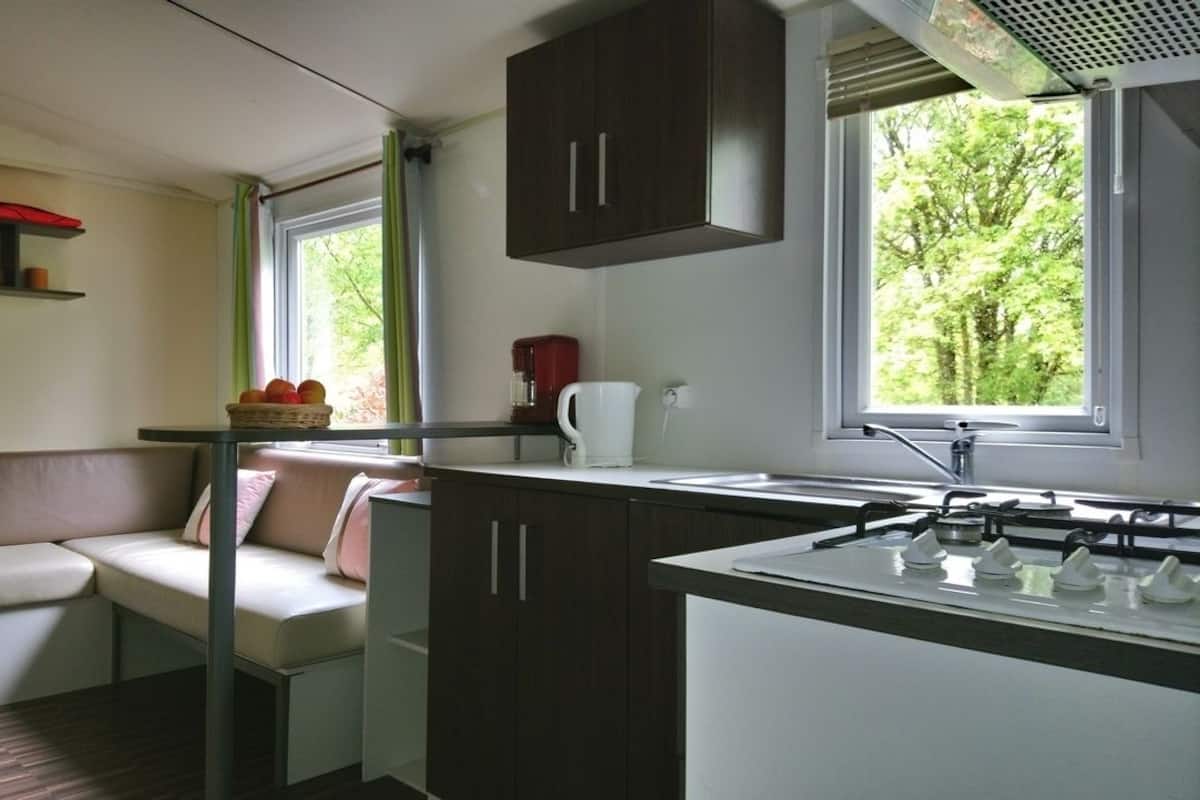 Une cuisine de taille réduite, équipée d’une cuisinière à gaz, d’un évier et d’une fenêtre donnant sur les arbres.