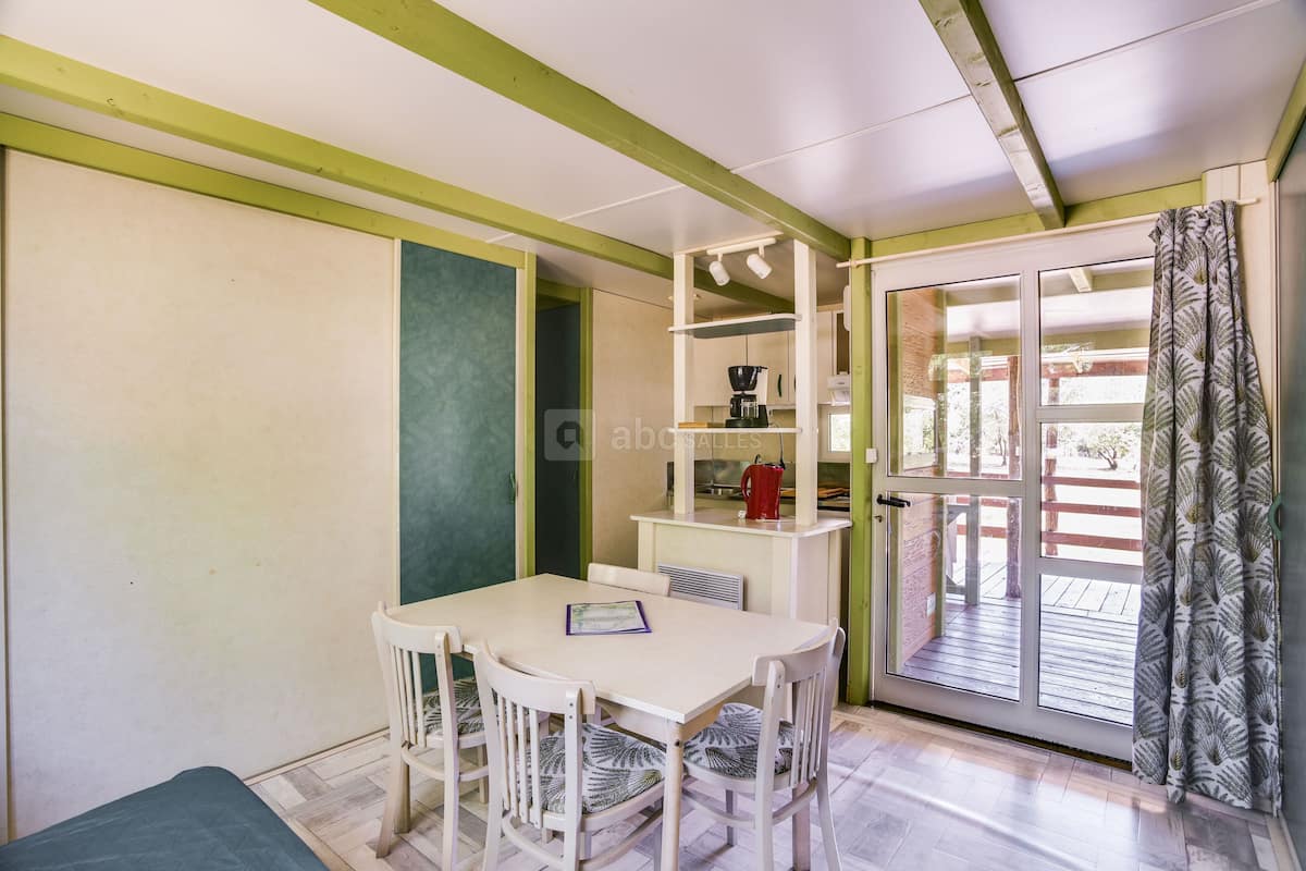 Un espace repas comprenant une table et des chaises, une machine à café et une vue sur une terrasse extérieure, accessible par une porte vitrée.